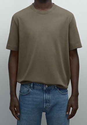 Mężczyzna ubrany w oliwkowozielony t-shirt z krótkim rękawem i okrągłym dekoltem, włożony w niebieskie dżinsy na jednolitym tle.