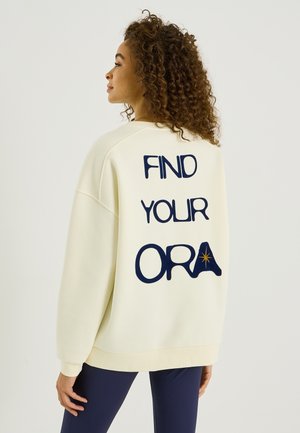 Sweat-shirt crème avec lettrage bleu marine au dos indiquant "FIND YOUR ORA" et une petite étoile. Présente une coupe décontractée et des poignets côtelés.