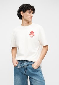 Λευκό βαμβακερό μπλουζάκι με στρογγυλή λαιμόκοψη και κοντά μανίκια, με κόκκινο γραφικό κείμενο στο στήθος. Συνδυάζεται με μπλε τζιν παντελόνι.