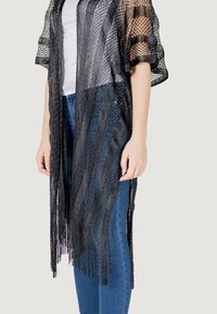 Copricostume in stile kimono nero con rete aperta, design a righe e orlo frangiato. Indossato su una maglietta bianca e abbinato a jeans blu.