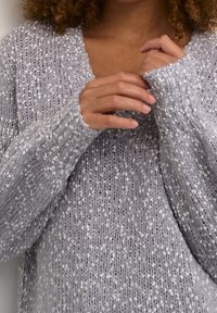 Pull en tricot gris clair avec un col en V, des poignets côtelés et une texture mouchetée. Gros plan sur des mains tenant le tissu.