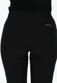 Zwarte broek van een gladde, rekbare stof. Bevat twee achterzakken en een logo-label. De tailleband zit comfortabel boven de heupen.
