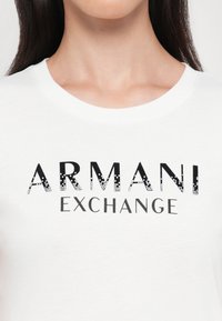 Balts kokvilnas T-krekls ar "ARMANI EXCHANGE" logo biezā melnā krāsā, ar gradienta tekstūru un smalkiem spīduma akcentiem.