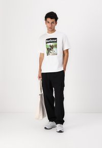 Tricou alb din bumbac cu un imprimeu grafic, purtat împreună cu pantaloni cargo negri, adidași albi și o geantă de pânză ușoară.