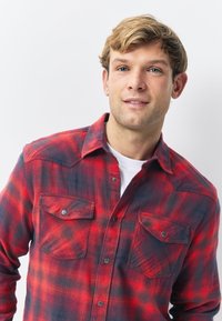 Cămașă de flanel în carouri roșu și negru, cu design cu nasturi, având două buzunare la piept și închidere cu nasturi de tip capse. Material moale și texturat.