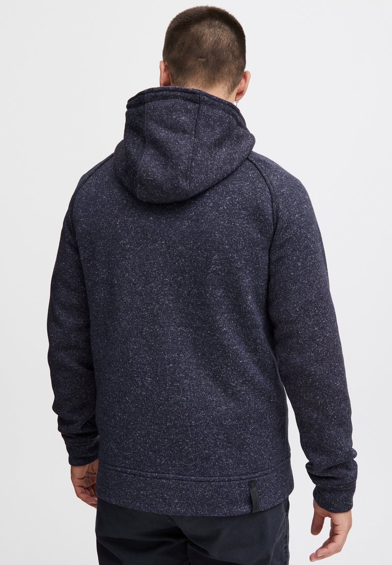 Homme portant un sweat à capuche bleu foncé moucheté, vu de dos sur un fond clair uni.