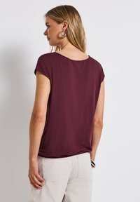 Burgunderfarbendes T-Shirt mit kurzen Ärmeln und rundem Ausschnitt, aus weichem Stoff, lockerer Passform und mit einer Nahtverzierung entlang dem Saum. Kombiniert mit hellen Hosen.