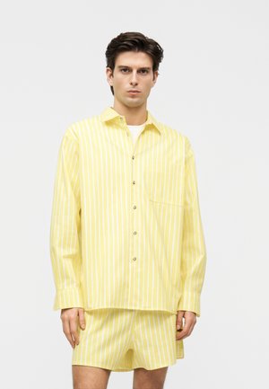 Jauns vīrietis, vilcis dzeltenu un baltu vertikālu svītru garroku kreklu un atbilstošus šortus, stāv pie vienkārša gaiša fona.