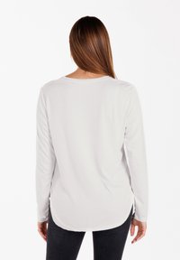 Langärmliges weißes Hemd mit rundem Halsausschnitt, weichem Stoff, seitlichen Schlitzen und einem abgerundeten Saum hinten, kombiniert mit dunklen Jeans.