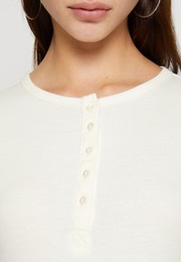 Vitt ribbat linne med rund halsringning och en knappslå med sex klara knappar, vilket framhäver en slät textur.