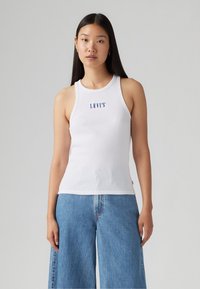 Baltas įstrižinis tank topas su apvaliu kaklu ir mėlynu "LEVI'S" logotipu ant krūtinės, derinamas su šviesiai mėlynomis plačiomis kelnėmis.