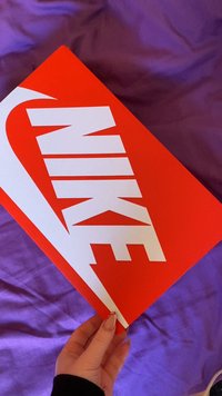 Sarkana Nike kurpju kaste ar lielu baltu swoosh logo un izteiksmīgu balto "NIKE" tekstu, turēta pret purpura auduma fonu.