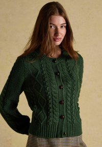 Zielony, dziergany kardigan z wzorami w warkocze, ściągaczem na dole i brązowymi guzikami. Tekstura jest gruba, z okrągłym dekoltem i dopasowanymi rękawami.