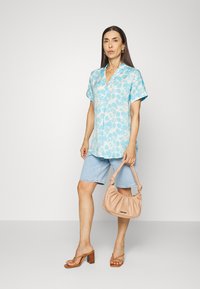 Camisa azul clara de manga curta com padrão floral, combinada com calções de ganga claros. Os acessórios incluem uma mala pêssego e sandálias de tacão baixo em tom bege.