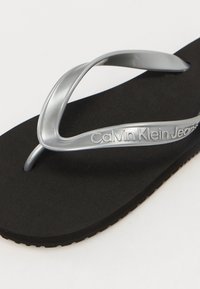 Svart flip-flop med en teksturert gummisåle og glansfull sølvrem. "Calvin Klein Jeans" er preget på remmen.
