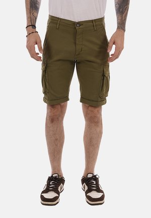 Uomo che indossa pantaloncini cargo verde oliva con tasche anteriori e sneakers bianche e marroni, in piedi davanti a uno sfondo neutro.