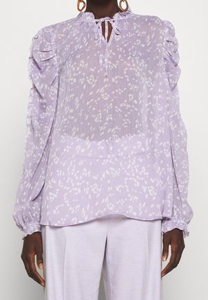 Personne portant un chemisier léger violet clair transparent avec des taches abstraites blanches, des manches à volants et un col noué, assorti à un pantalon de couleur claire.
