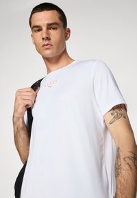 Hvid atletisk t-shirt med et HYROX-logo i orange, korte ærmer, rund halsudskæring og en let, glat tekstur.