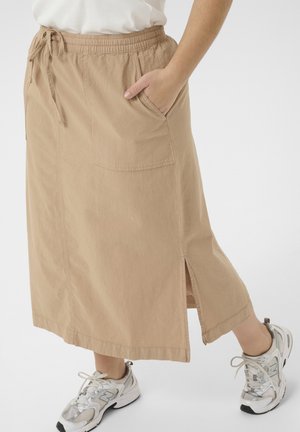 Beige midirok met voorzak en zijsplit, gedragen met witte sneakers en een ingestopt wit topje, hand rustend in de zak.