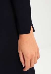 Hand liegt entspannt an der Seite und trägt ein langärmliges, schwarzes Strickkleidungsstück mit einem kleinen Schlitz am Bündchen vor neutralem Hintergrund.