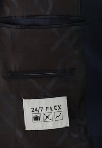 Schwarzer Stoff mit glatter Textur und dezentem, geometrischem Muster, versehen mit einem eingenähten Etikett "24/7 FLEX" und drei Symbolen, die die Funktionalität anzeigen.