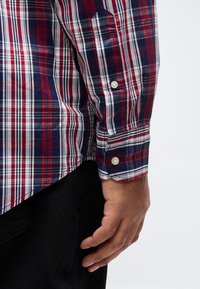 Χέρι άνδρα που ακουμπάει δίπλα στο ισχίο, φοράει μακρυμάνικο πουκάμισο καρό σε κό κκινο, άσπρο και ναυτικό μπλε με μανσέτα με κουμπιά και μαύρο παντελόνι.