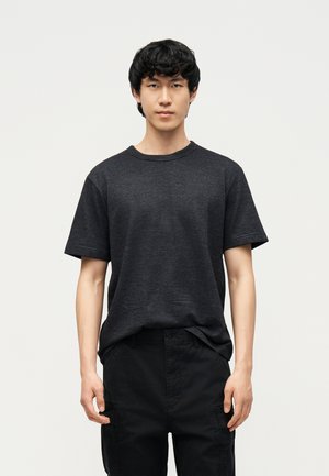 Ung mand med kort sort hår iført en mørkegrå, stribet kortærmet skjorte og sorte bukser, stående foran en ensfarvet lys baggrund.