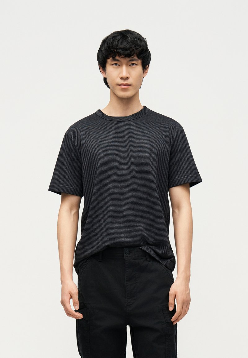 Νέος άνδρας με κοντά μαύρα μαλλιά, φοράει σκούρο γκρι ριγωτό κοντομάνικο πουκάμισο και μαύρο παντελόνι, στέκεται μπροστά σε απλό ανοιχτό φόντο.