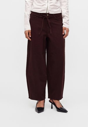 Vrouw draagt donkerbruine, wijde broek met een koord in de taille en zwarte pumps met een puntige neus op een witte achtergrond.