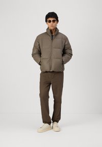 Brun pufferjakke med høj krave, lynlås foran og vandrette syninger. Sammen med brune bukser og off-white sneakers. Teksturstoff.