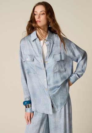 Jeune femme aux longs cheveux portant une chemise en denim clair oversize et un pantalon, accessoirisée de bracelets bleus et argentés, posant devant un fond uni.