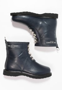Las botas de goma náuticas hasta el tobillo presentan una suela negra texturizada, ojales plateados y una etiqueta con la marca en el costado, con un lazo para un fácil calzado.