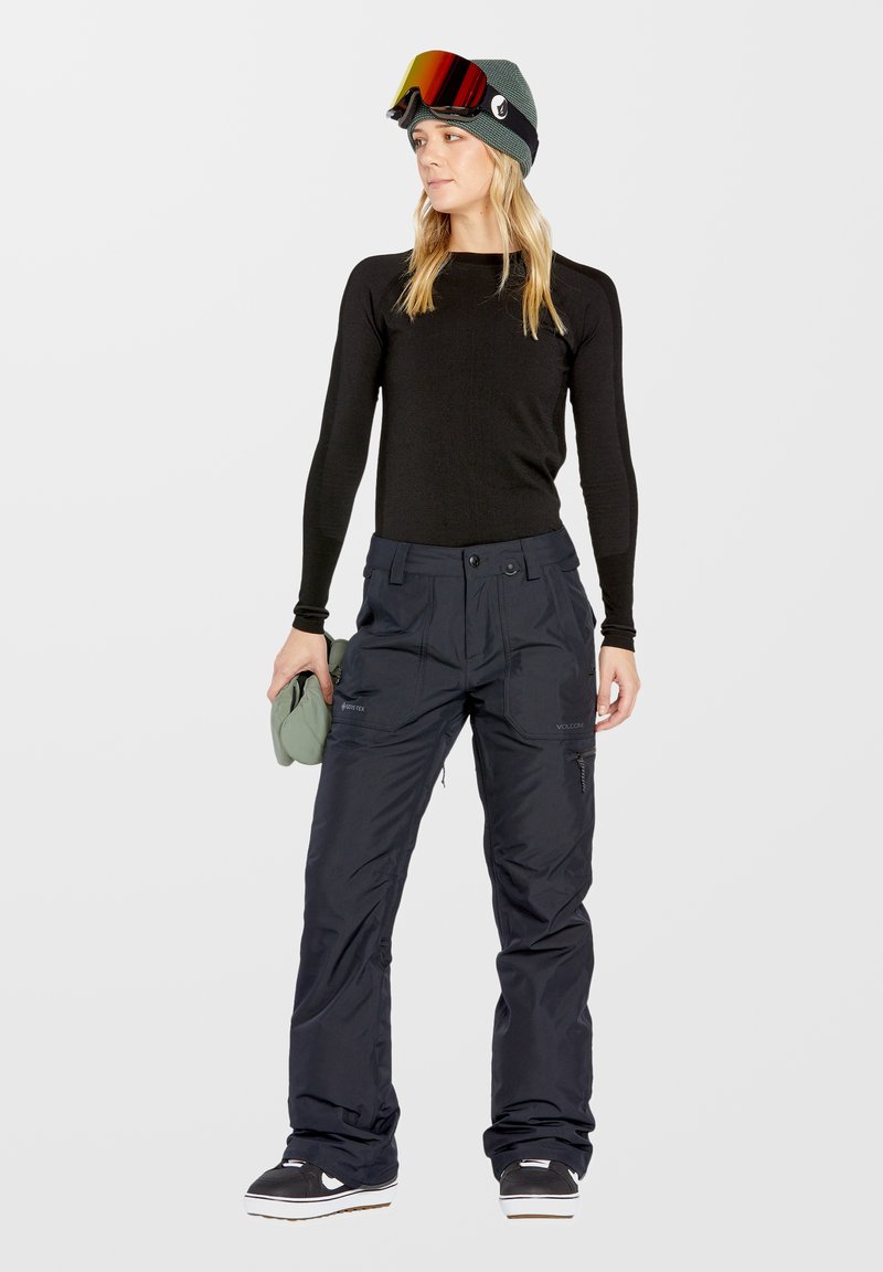 Top à manches longues noir, pantalon de neige bleu marine et bonnet vert avec lunettes de protection. Le pantalon présente plusieurs poches et un design à coupe décontractée.