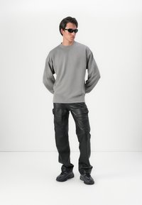 Felpa grigia oversized con un piccolo logo rosso, abbinata a pantaloni cargo in finta pelle nera e sneakers nere. Modella che indossa occhiali da sole.