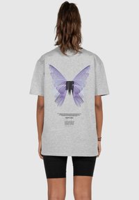 Graues Baumwoll-T-Shirt mit einem großen, detaillierten lila Schmetterlingsmotiv auf dem Rücken, versehen mit schwarzem Text und zusätzlichen Grafikelementen.