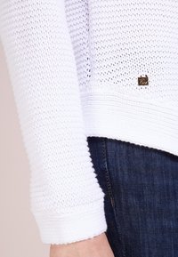 Pull en tricot blanc avec un motif texturé, doté de poignets côtelés et d'un petit logo doré près de l'ourlet. Porté avec un jean en denim foncé.