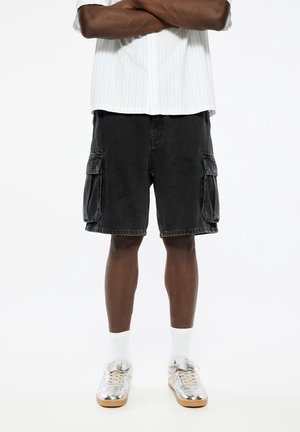 Schwarzjeans Cargo-Shorts mit Seitentaschen, kombiniert mit silbernen Sneakern und weißen Socken. Darüber ein einfaches kurzärmeliges Hemd mit Streifen.