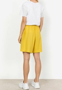 Biała koszula z koronkowymi rękawami, zestawiona z żółtymi szortami z elastycznym pasem, noszona z białymi trampkami na neutralnym tle.