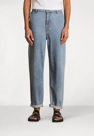 Personne portant un jean en denim bleu clair retroussé, un haut blanc et des sandales noires à bouts ouverts, debout devant un fond uni.
