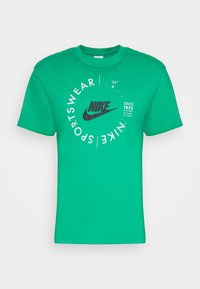Vihreä puuvillainen t-paita lyhyillä hihoilla, jossa on musta Nike-logo ja pyöreitä graafisia elementtejä, mukaan lukien "Sportswear" ja "Since 1972."