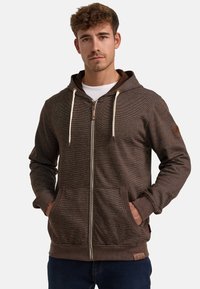 Brown zip-up hoodie with a textured surface, adjustable drawstrings, front pockets, and a logo patch on the sleeve.