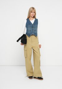 Δερμάτινο γιλέκο με πέντε κουμπιά, ανοιχτό μπλε, φορεμένο πάνω από ένα λευκό πουκάμισο με μακριά μανίκια. Καφέ cargo παντελόνι με τσέπες. Μαύρη τσάντα κρατημένη σε ένα χέρι.