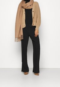 Cardigan beige lavorato a maglia con grandi bottoni, top nero, pantaloni neri e scarpe con tacco color cuoio; abbinato a una sciarpa beige coordinata.