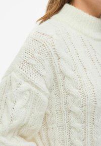 Pulover s kabelskim pletenjem iz mehkega off-white preje, z visokim ovratnikom, teksturiranimi vzorci in vidnimi šivi, ki ustvarjajo prijeten videz.