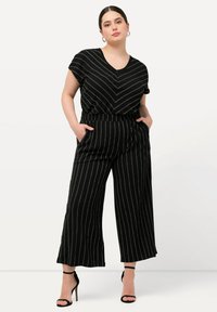 Combinaison noire rayée avec un col en V, des manches courtes et une taille cintrée. Présente un pantalon large et des poches latérales. Texture lisse.