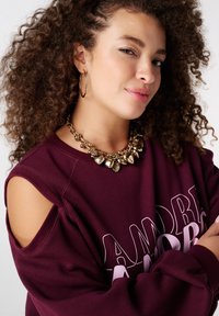 Sweat-shirt bordeaux avec design à épaules découpées, avec de grandes lettres blanches. Collier chunky doré avec des breloques en forme de cœur et de coquillage.