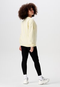 Lichtgele sweatshirt met geribbelde manchetten, gecombineerd met zwarte leggings. Het model draagt witte sneakers en heeft krullend haar.
