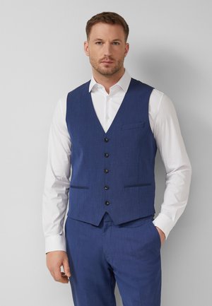 Mężczyzna ubrany w niebieską kamizelkę zapinaną na guziki i dopasowane spodnie, na białej koszuli z długim rękawem, stojący na tle jednolitego jasnego tła.