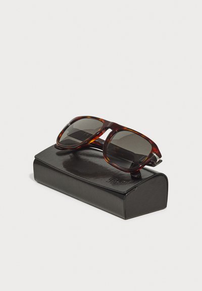 Tortoiseshell sunglasses with dark lenses, placed on a black leather case. Case shows a curved shape and embossed logo detail.