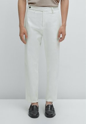 Pantaloni bianchi con una vestibilità sartoriale, leggero lucido, tasche laterali e chiusura a bottone. Abbinati a mocassini neri. Tessuto dalla consistenza liscia.